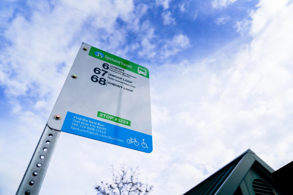 A Spokane Transit bus stop sign with route numbers 66 (EWU), 67 (Three Loop), and 68 (Cheney Loop) under a partly cloudy sky. The sign includes the stop number 3224 and contact information for real-time bus schedules.