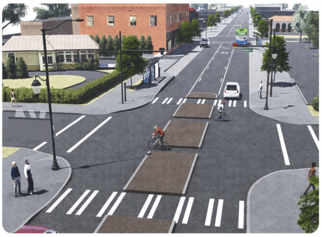 A city street with two bicyclists crossing at a crosswalk. Several cars travel on the street, and pedestrians wait at the corner. The area is lined with buildings, trees, and greenery, featuring bike lanes and designated crosswalks.