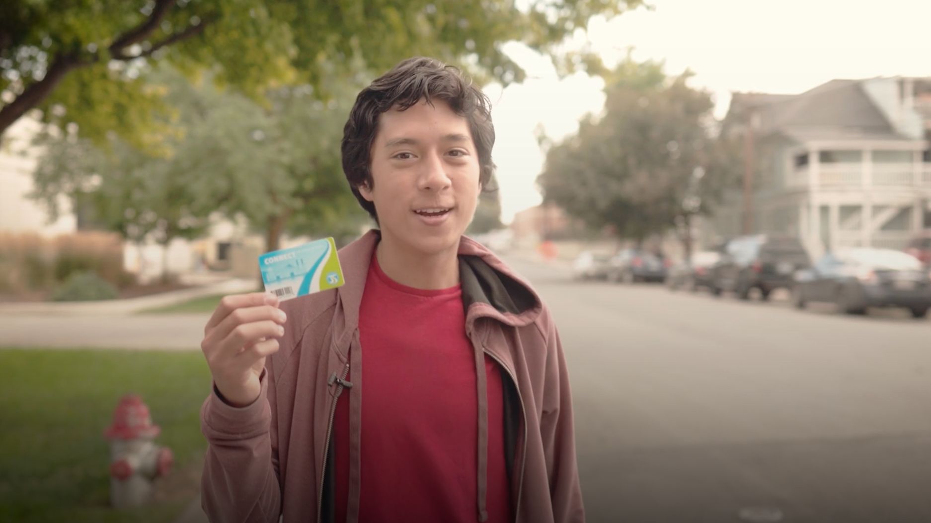 A person in a red shirt and brown jacket stands outdoors on a residential street, holding up a blue and green card that says Zero Fare for Youth. Trees and parked cars line the street in the background, and houses are visible on both sides. The person is smiling and looking at the camera.