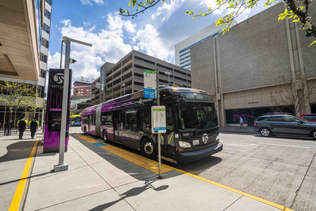A modern city bus with "City Line" branding is at a bus stop on an urban street. The surrounding area features tall buildings, a bus shelter, and people walking. The sky is partly cloudy, and there is a signpost with bus route information near the bus.