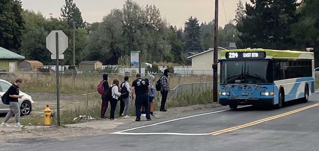 A group of people wait at a bus stop on the sidewalk next to a road. A blue and white bus with route number 29A and a sign reading "East 17th" approaches the stop.