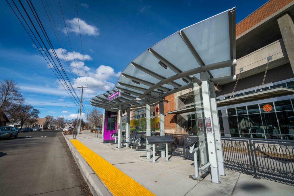 A modern bus stop with a glass canopy, silver metal benches, bike racks, and a yellow tactile paving strip along the sidewalk. A street and power lines are on the left, and buildings are visible in the background under a blue sky with clouds.