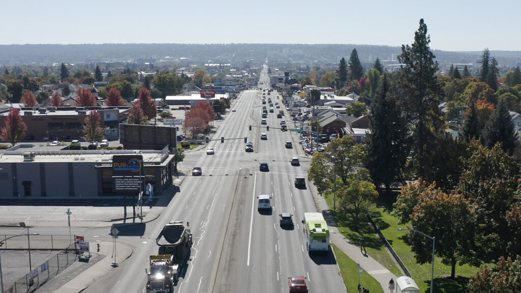 A wide aerial view of a busy urban street lined with trees, vehicles, and buildings. The road stretches into the distance under a clear sky, with traffic moving in both directions. Fall foliage is evident with trees displaying a variety of autumn colors.