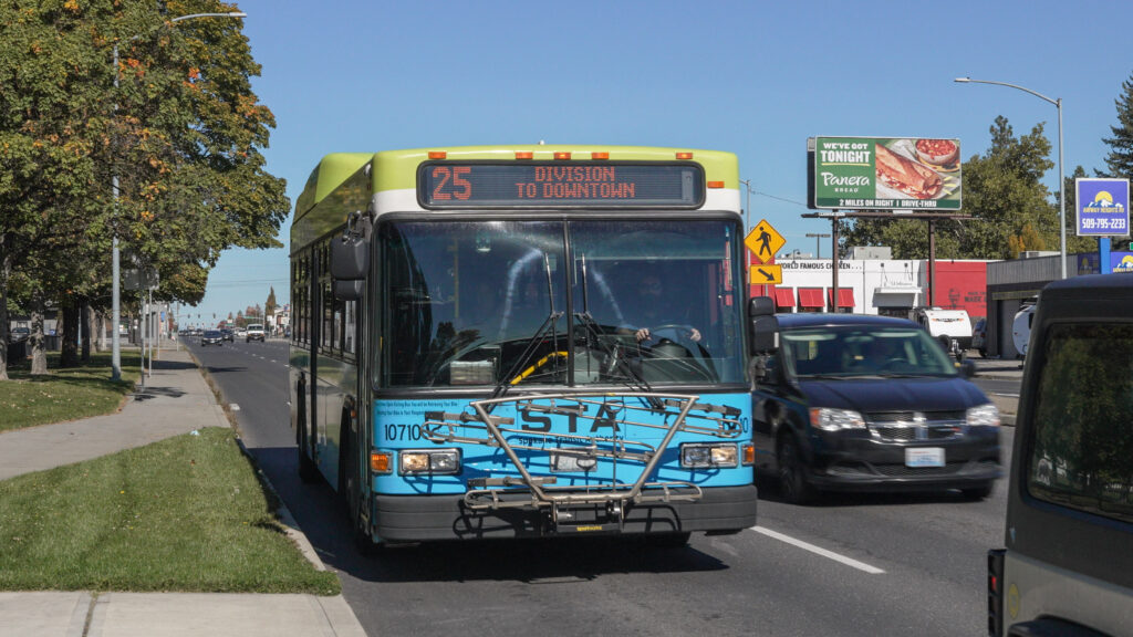 A city bus with route number 25 and "Division to Downtown" displayed on the front is driving on a street with other vehicles. There are trees and a billboard with an advertisement along the roadside. The sky is clear and blue.
