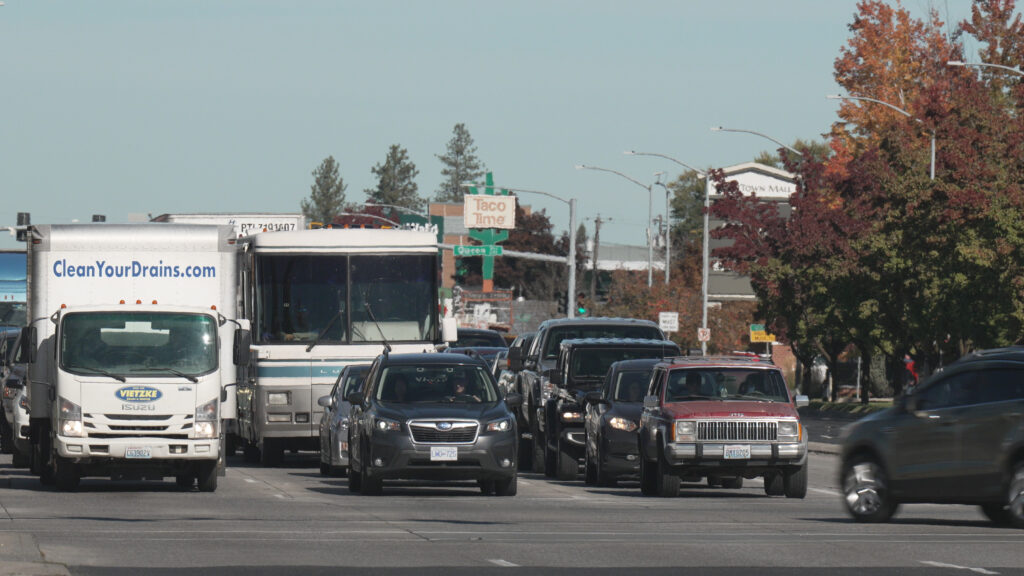 A busy urban street with multiple lanes of vehicles, including cars, trucks, and an RV, all waiting at a traffic light. Trees with autumn foliage are visible on the right side, and a "Taco Time" sign is in the background. The sky is clear.