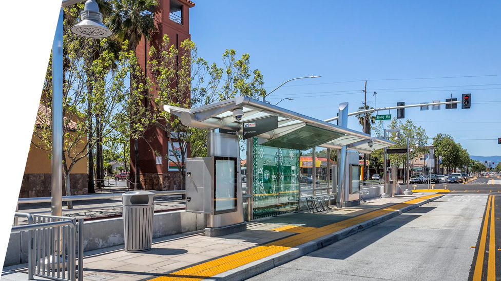 A modern bus stop in Santa Clara with a glass shelter, seating, and digital information displays, located on a sunny urban street with clear blue skies, traffic lights, and lush green trees.