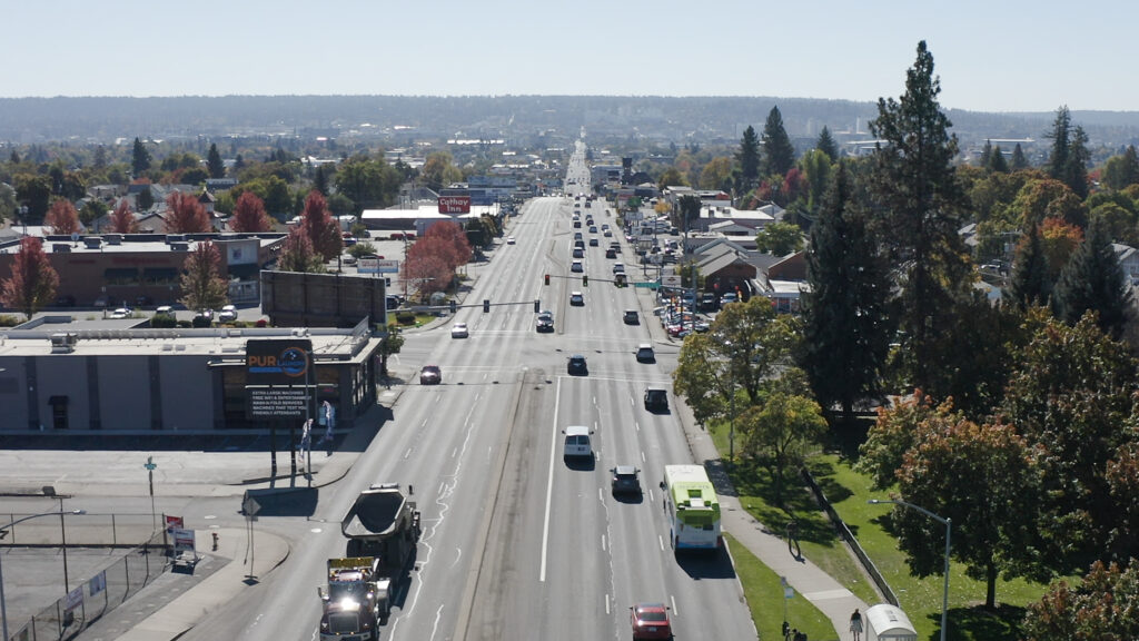 Aerial view of a multi-lane road in Spokane with transit vehicles, surrounded by buildings, trees with autumn foliage, and distant hills under a clear sky.