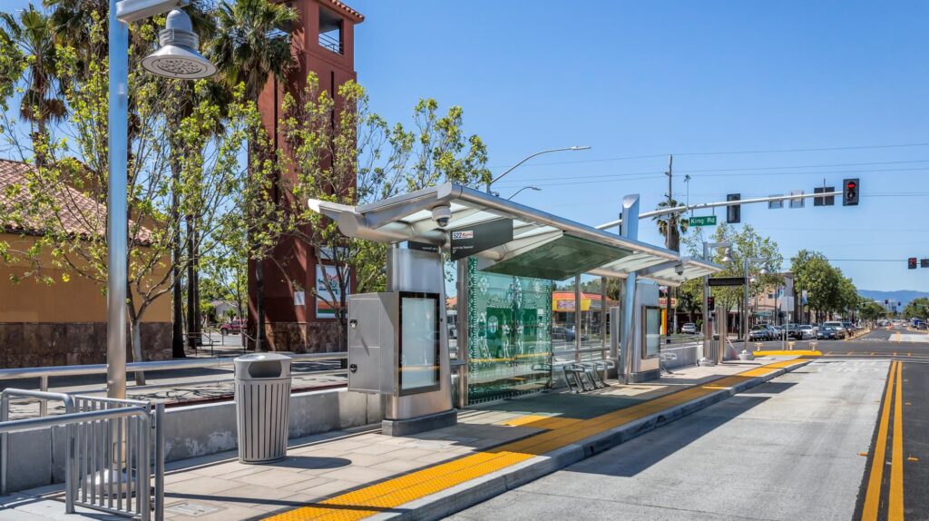 A modern bus stop in Santa Clara with a glass shelter, metal benches, and digital display, located on a sunny street with clear blue skies and a pedestrian crossing in the background.