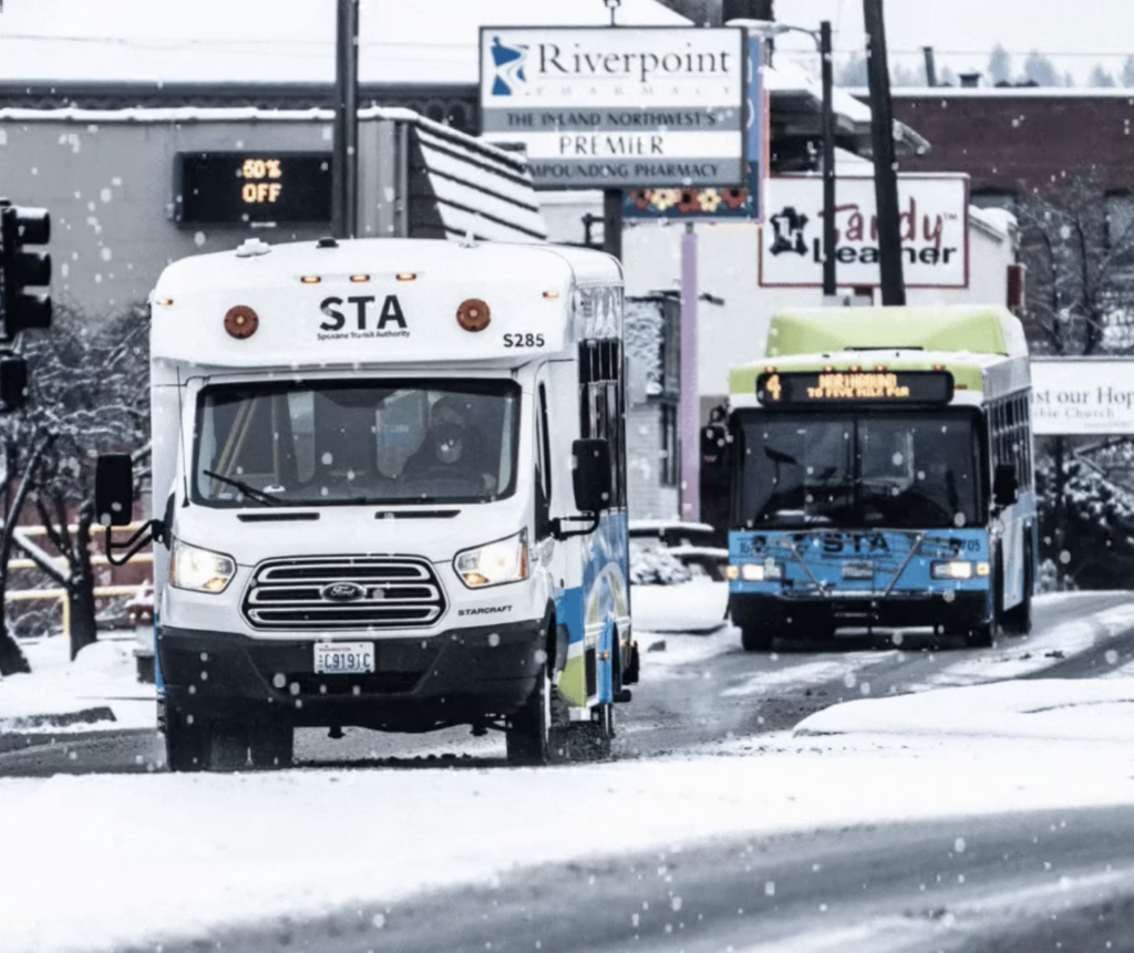 Two STA buses drive on a snowy street in a city. The bus in front is smaller and white, while the bus behind is larger and blue-green. Snow covers the ground and rooftops, and various business signs are visible in the background.