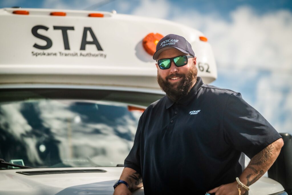 A bearded man wearing sunglasses, a cap, and a polo shirt with the "Spokane Transit" logo stands confidently beside a Spokane Transit Authority bus. With employee benefits that include job security and health perks, the sky is partly cloudy in the background.