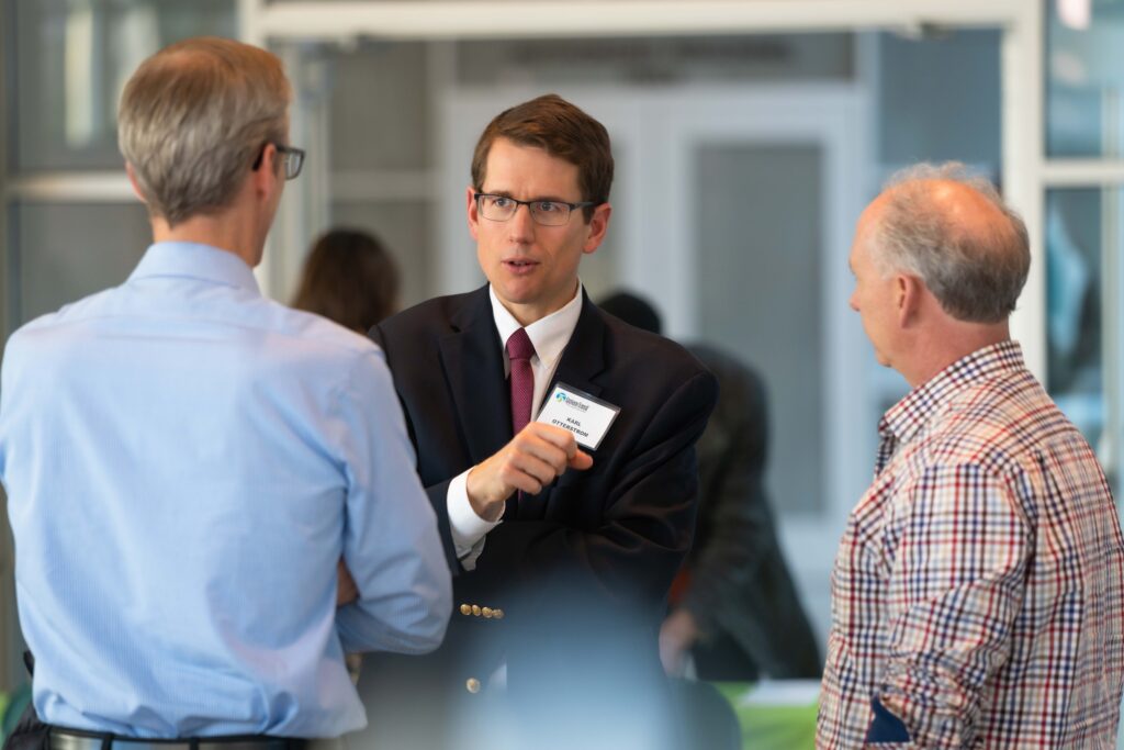 A man in a suit and tie with a name tag speaks animatedly to two other men, one in a light blue shirt and the other in a checkered shirt. They are in a bright indoor setting, possibly at a professional event or seminar.