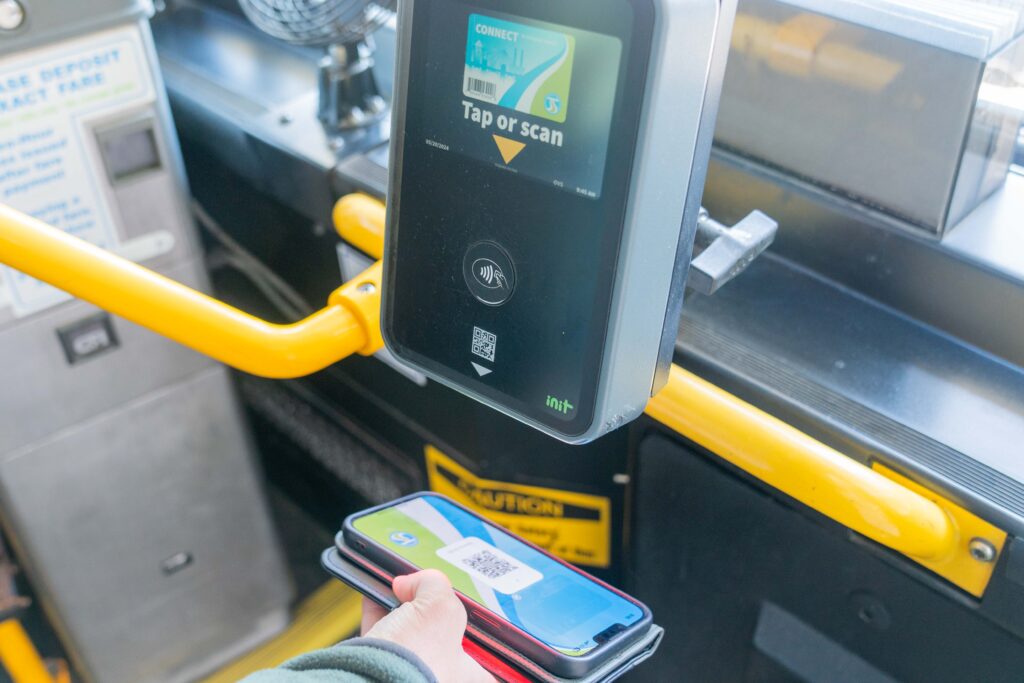 A person holds a smartphone with a QR code displayed on its screen near a contactless payment terminal on a public bus, ready to tap or scan for fare payment. Yellow handrails and safety information are visible in the background.