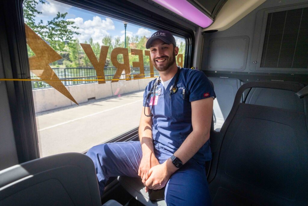 A person wearing medical scrubs and a cap sits inside a vehicle, smiling at the camera. They have a stethoscope around their neck and are positioned next to a window on a sunny day. The window has partially visible writing on it.