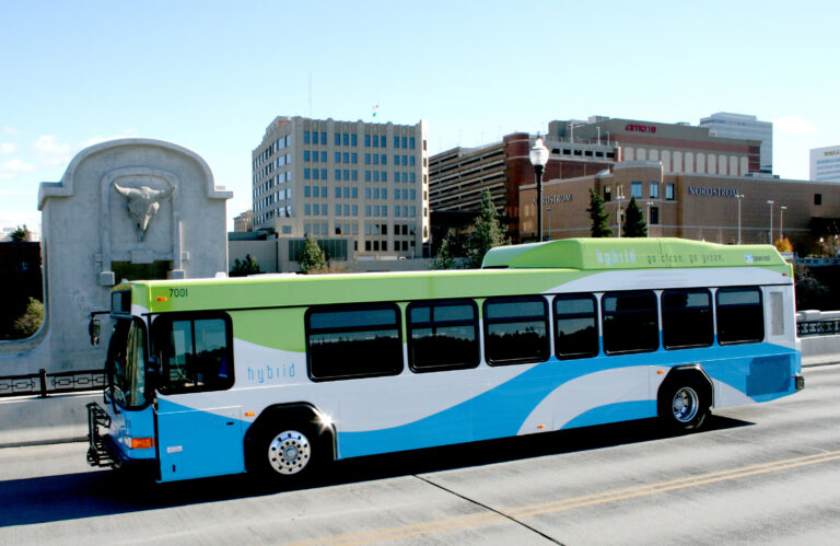 A hybrid bus on a bridge with city buildings and a bull sculpture in the background on a clear day.
