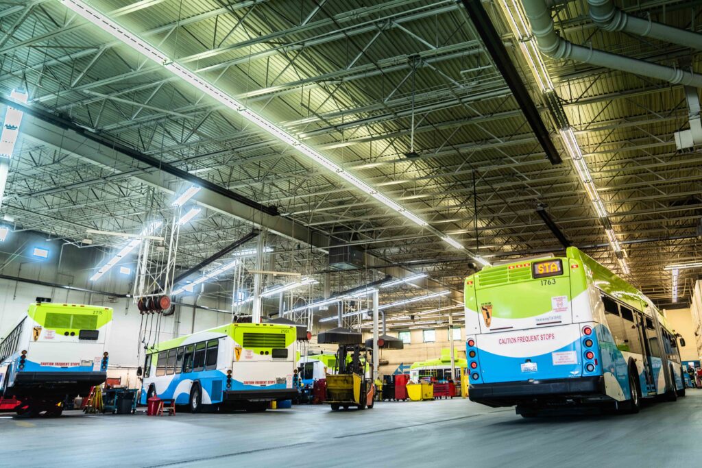 A large, well-lit bus depot with several buses parked inside undergoing maintenance and repairs.