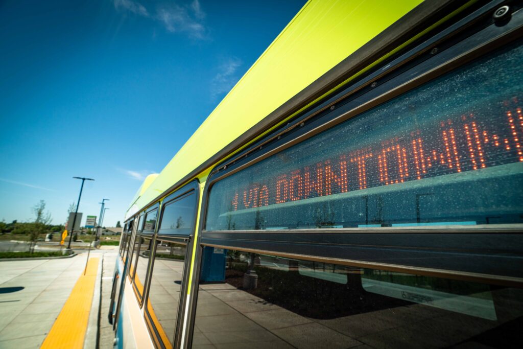 Close-up of a bus with a digital sign reading "4 VIA DOWNTOWN" at a sunny outdoor bus stop.