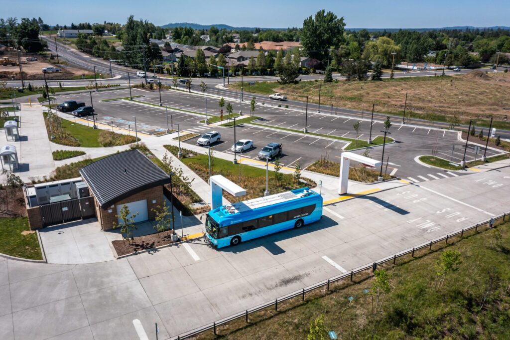 A blue bus at an empty parking lot near intersection with trees, grass, and buildings in the background on a sunny day.