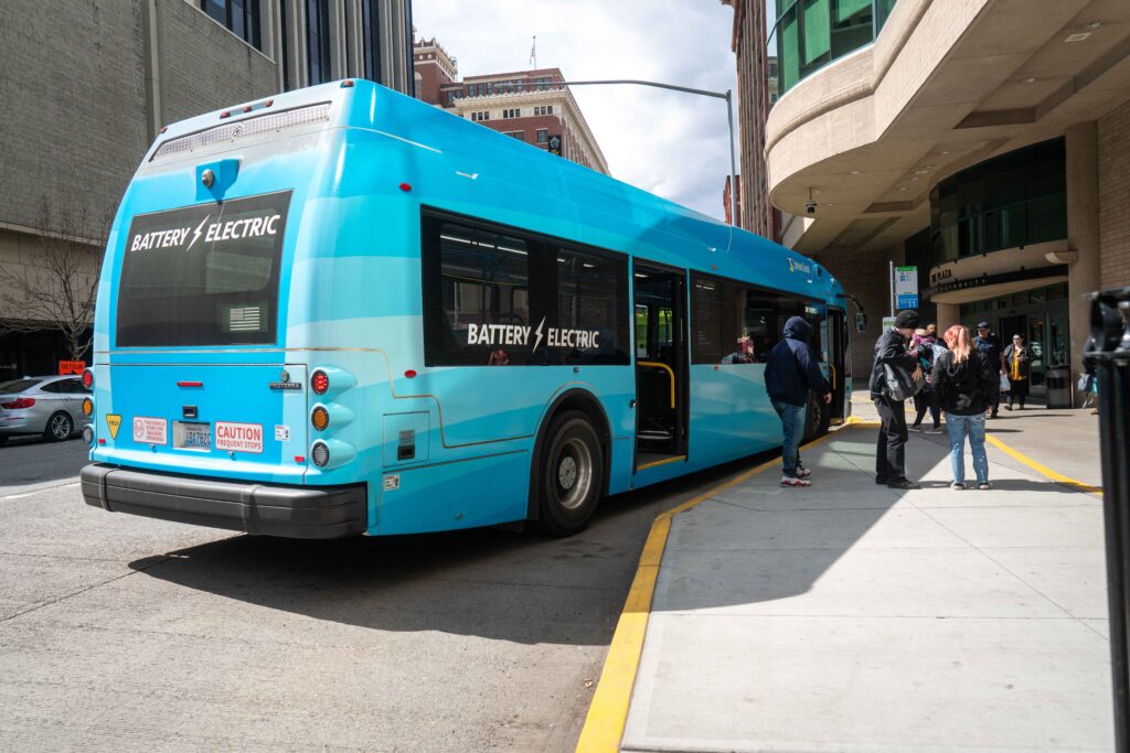 A blue battery-electric bus is parked at a stop with several people waiting to board.