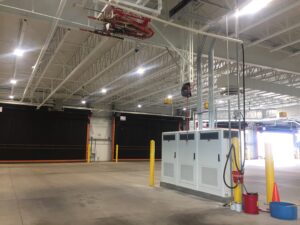 Spacious industrial warehouse with high ceilings, bright lighting, and various equipment including red fire safety gear.