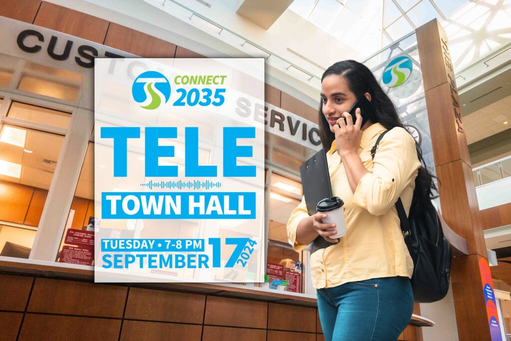 A woman holding coffee and a tablet talks on the phone next to a sign promoting a September 17 Tele Town Hall event.