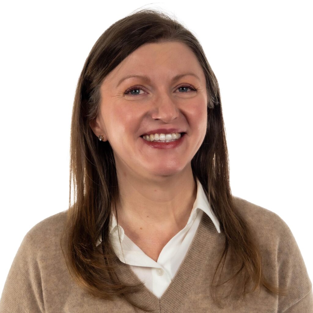 Smiling woman with long brown hair in a beige sweater over a white shirt against a white background.