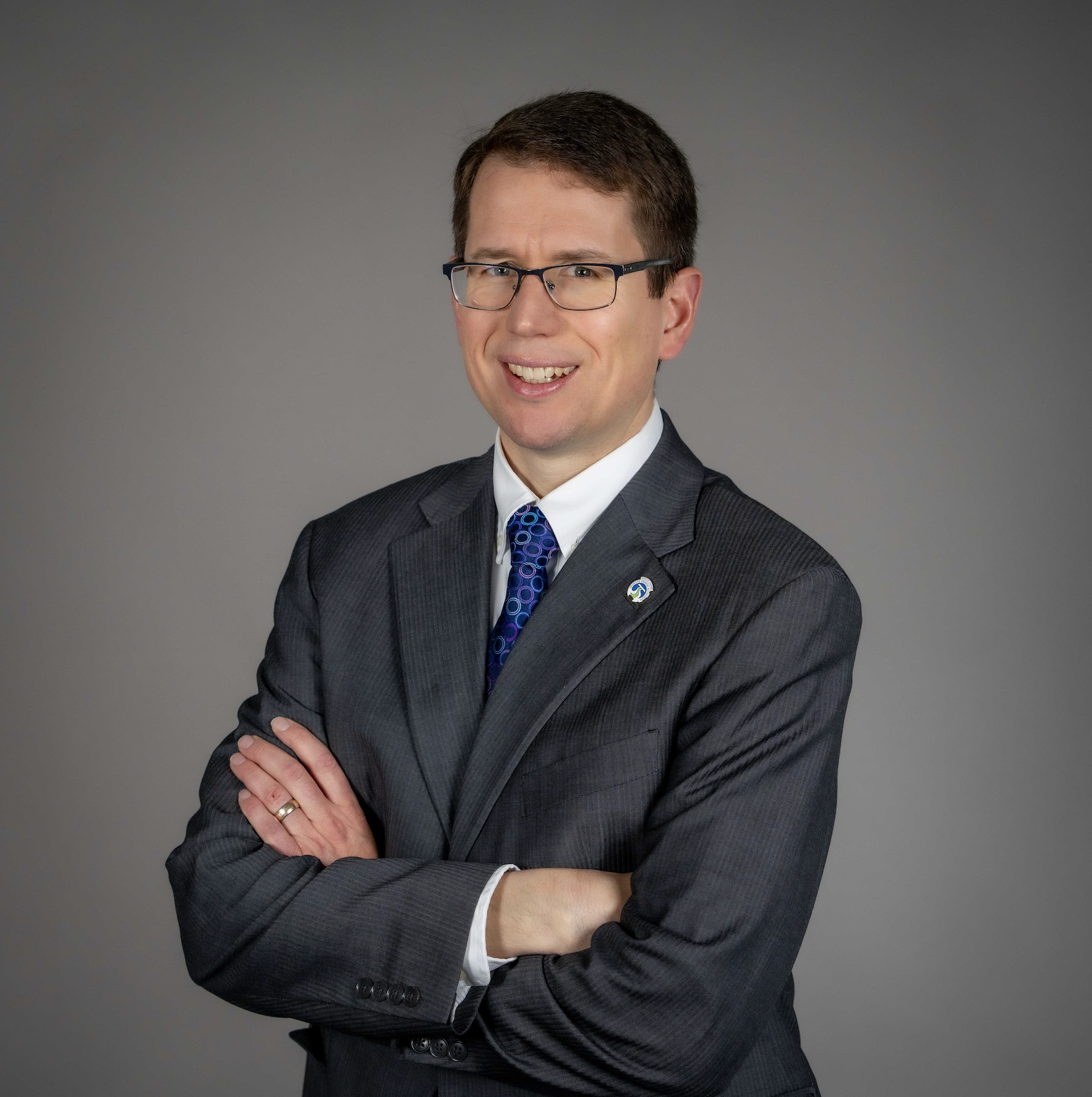 Person in a gray suit with glasses and a blue tie, smiling with arms crossed against a plain background.