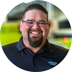 Smiling man wearing glasses and a black "Spokane Transit" shirt in a brightly lit indoor setting.