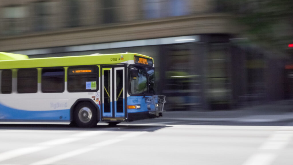 Blurred image of a moving city bus with a green and blue color scheme on a street.