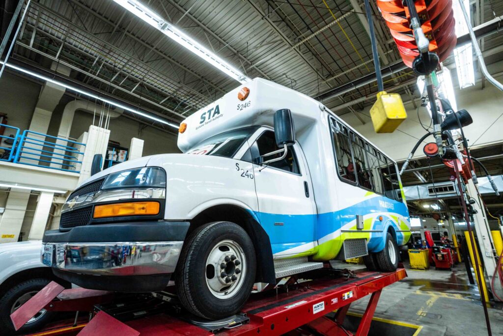 A white and blue bus on a service lift in a garage, surrounded by various tools and equipment.