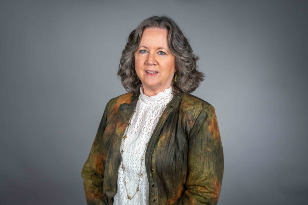 STA's Clerk of the Authority Dana Infalt, wearing a colorful jacket and white lace blouse and standing against a gray background.
