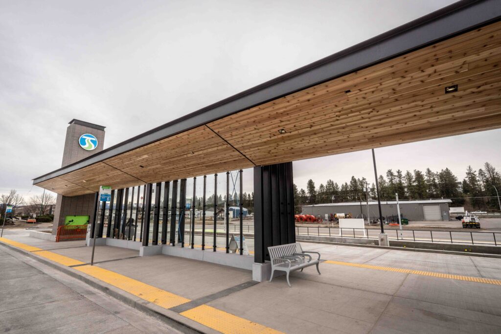 Bus station with a wooden roof, a bench, and a sign on the wall. Overcast sky and trees in the background.