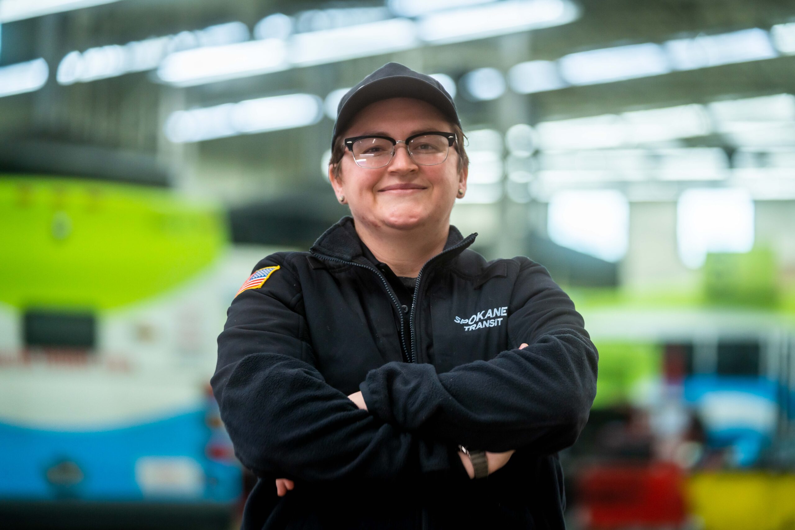 STA bus in indoor transit facility with a smiling person wearing glasses, cap, and Spokane Transit jacket standing with arms crossed.