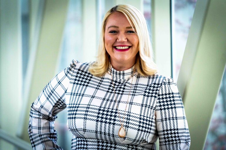 A woman with blonde hair in a black and white plaid dress smiles in front of large geometric windows.