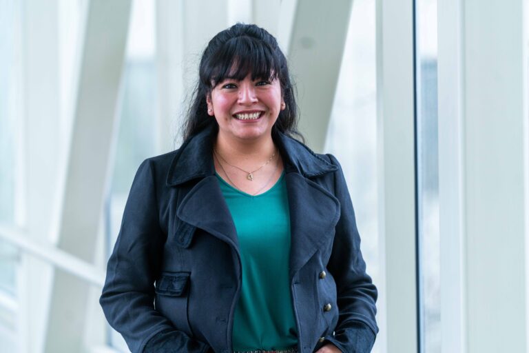 Yolanda Montes smiles indoors by large windows, wearing a green top and dark jacket.