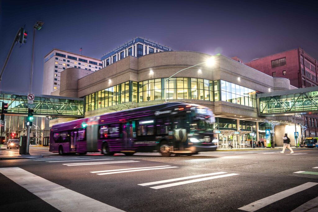 An STA bus drives past a modern transit station with skywalks at dusk; buildings and streetlights are in the background.