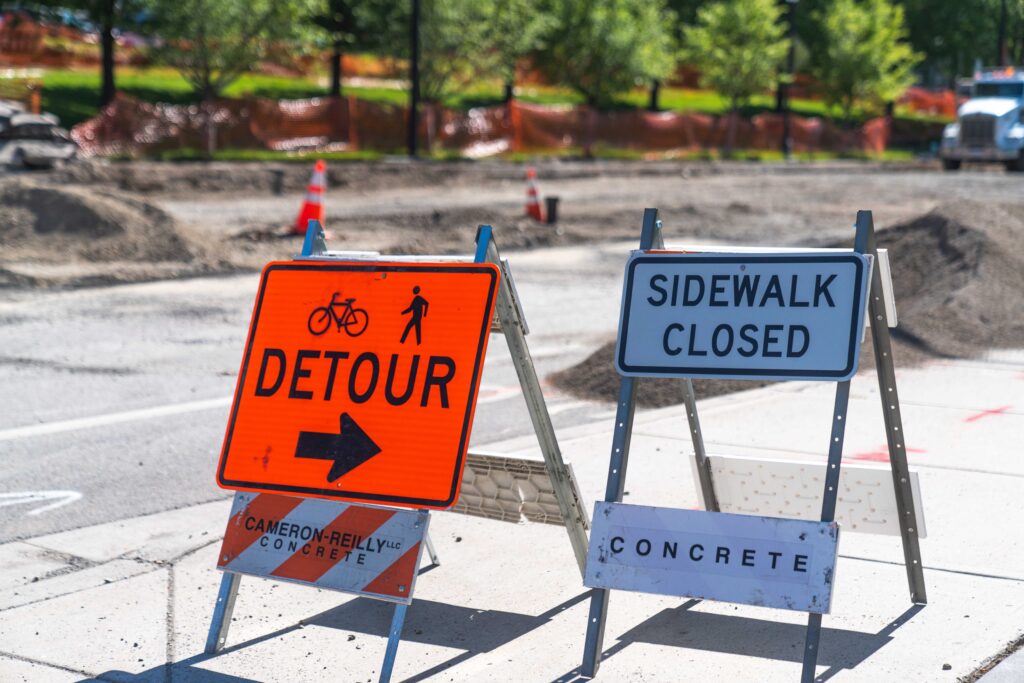 Orange detour sign with bike and pedestrian icons points right; "Sidewalk Closed" sign next to street construction area.