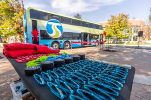 Promo items, including sunglasses and drink coasters made from recycled tires, on a table in front of a ribbon-adorned double-decker bus.