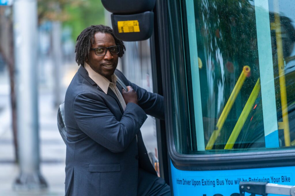 Man in suit and glasses steps onto a city bus, looking back and smiling.