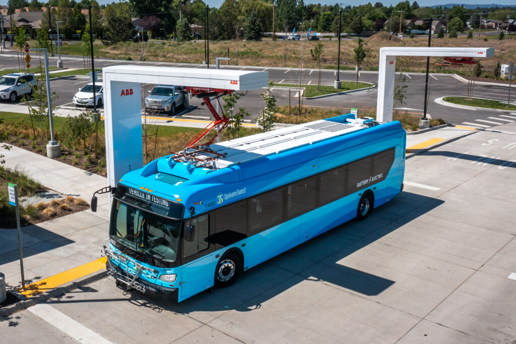 Blue electric bus charging at a station in a parking lot.