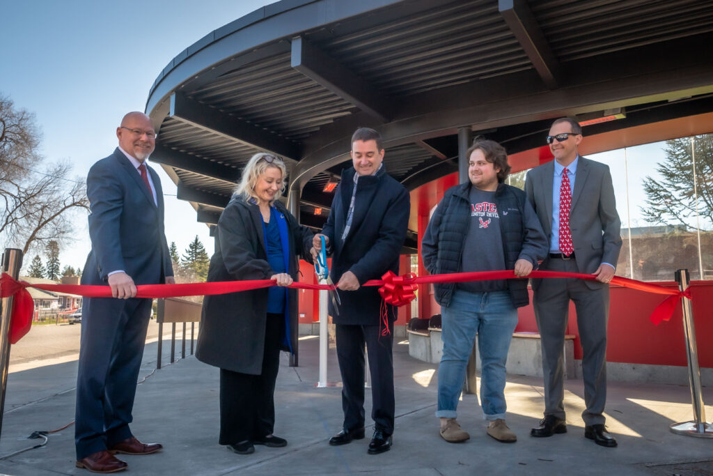 Five people cut a red ribbon in front of a building at a ribbon-cutting ceremony.