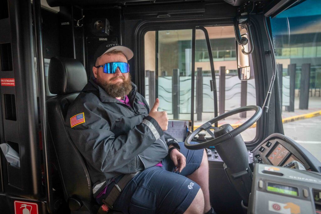 Bus driver with reflective sunglasses gives a thumbs up from the driver’s seat.