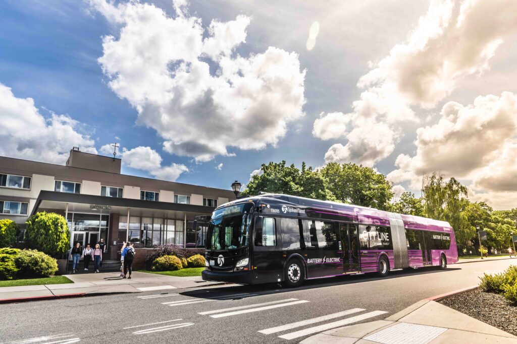 Purple articulated bus stopped by a modern building; a few people stand nearby.