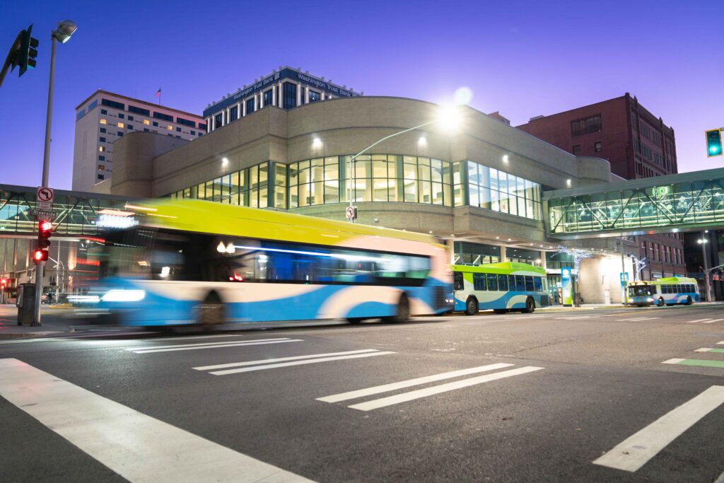 A city bus drives past a modern transit station at dusk; another bus sits nearby beneath a skybridge.
