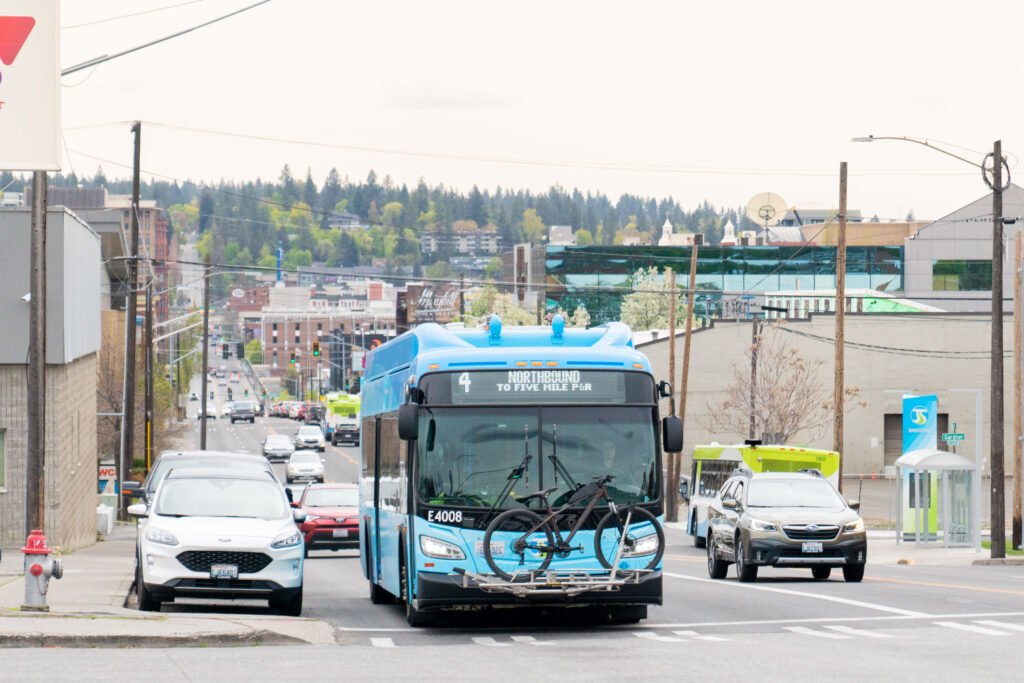 Blue city bus drives uphill, with other cars and urban buildings nearby.
