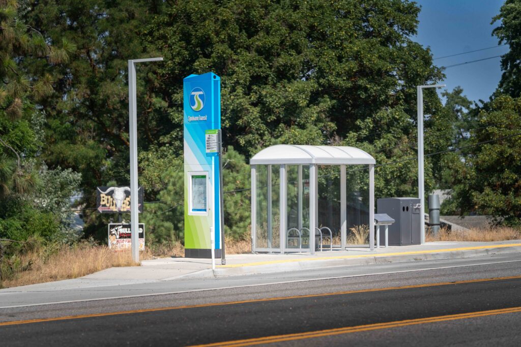 Modern bus stop with shelter and bike rack beside a road, surrounded by trees.