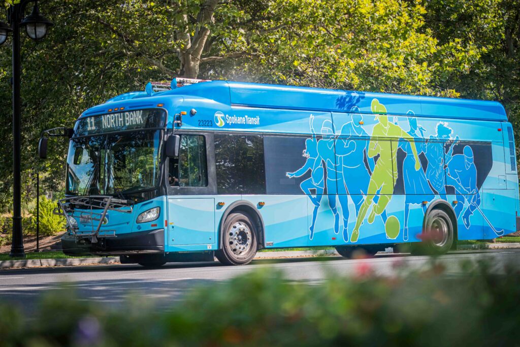 Blue Spokane Transit bus with sports-themed artwork on a street with green trees.