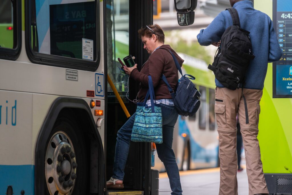 Woman boards city bus holding phone; man stands behind her in line at bus stop.