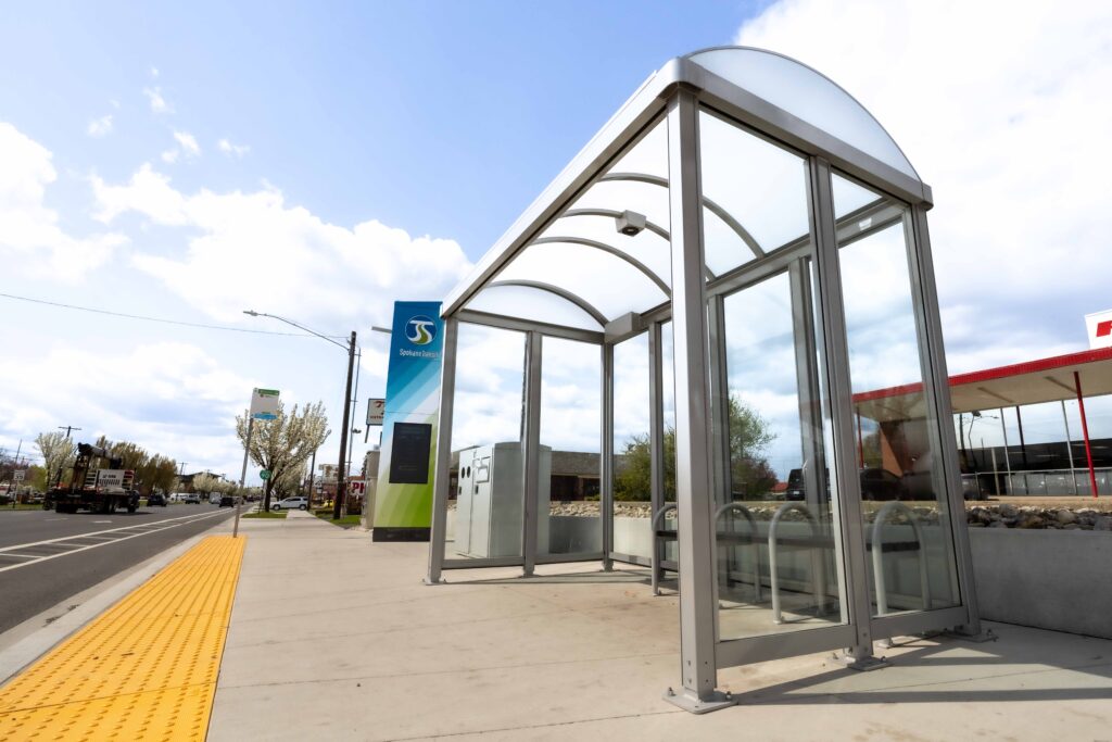 Glass bus shelter on sidewalk next to tactile yellow path along a road.