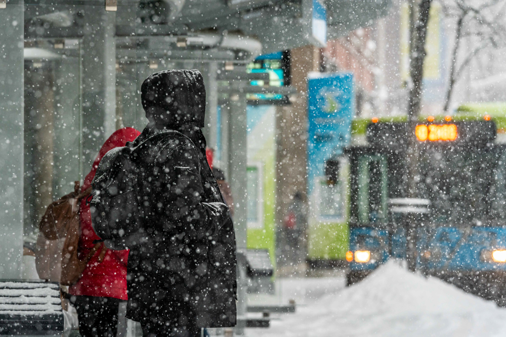 People wait at a snowy bus stop while a green bus approaches through heavy snowfall.