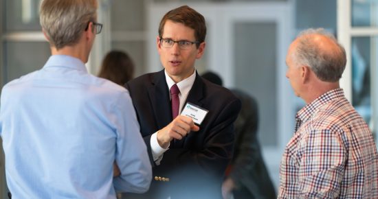 A man in a suit and tie with a name tag speaks animatedly to two other men, one in a light blue shirt and the other in a checkered shirt. They are in a bright indoor setting, possibly at a professional event or seminar.